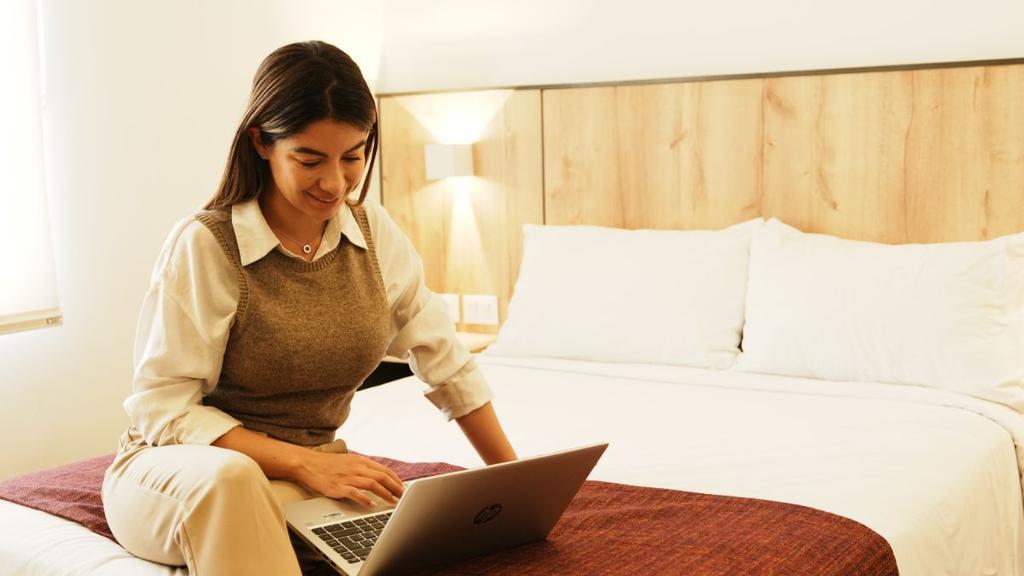 Persona usando laptop en cama con cabecera de madera y lámparas de pared
